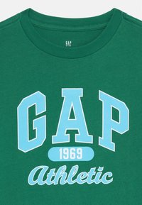 Grønn bomull T-skjorte med rund hals. Har blå grafisk tekst "GAP 1969 Athletic" i fet skrift på forsiden. Glatt tekstur.
