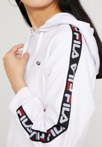 Felpa bianca con cappuccio e logo FILA ricamato in nero e rosso lungo le maniche. Presenta un cappuccio con coulisse e polsini a costine.