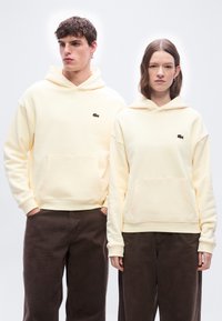 Νέος άνδρας και γυναίκα φορούν ταιριαστά εκρού φούτερ με κουκούλα και σκούρα καφέ παντελόνια, στέκονται ο ένας δίπλα στον άλλον μπροστά σε ένα απλό φωτεινό φόντο.