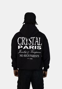 Felpa nera con grande testo bianco sul retro: "CRYSTAL PARIS Gioielli & Orologi NESSUN GENITORE RICCO." Il materiale sembra morbido.
