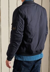 Navy bomber jacket with a quilted texture, ribbed cuffs, and waistband. Features a side zip pocket and layered denim shirt underneath.