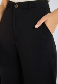 Main dans la poche avant de pantalons taille haute noirs avec un bouton et des passants de ceinture, associés à un haut blanc.