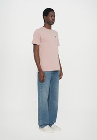 Helle rosa Baumwoll-T-Shirt mit kurzen Ärmeln, rundem Halsausschnitt und kleinem schwarzen Logo. Kombination mit weiten blauen Jeans und weißen Sneakers.
