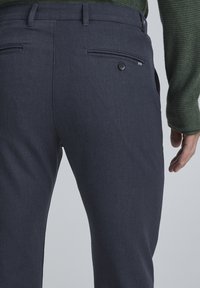 Mörkgrå byxor i twilltyg, med två bakfickor med knappstängning och en slim fit-design, bärs tillsammans med en grön strukturerad tröja.