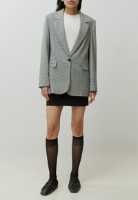 Grijze geruite blazer met geknipte revers en een enkele knoop, gecombineerd met een zwarte minirok en knielange zwarte doorschijnende sokken. Zwarte platte schoenen.