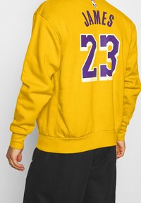 Sweat-shirt jaune avec poignets et ourlet côtelés. Présente "JAMES" et le numéro "23" en violet avec un contour blanc au dos. Design classique à enfiler.