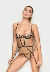 Mujer vistiendo lencería transparente nude con ribetes negros y patrones de estrellas, sosteniendo tirantes de liguero contra un fondo claro y sencillo.