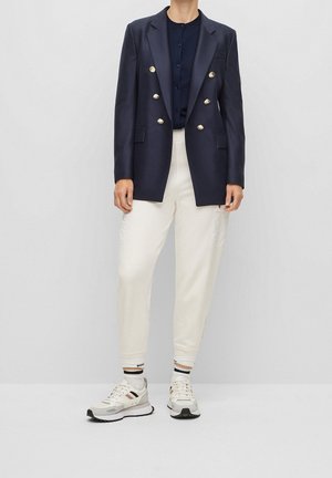 Blazer croisé bleu marine avec boutons dorés, associé à un pantalon blanc fuselé et des baskets gris-blanc, présente un design décontracté et élégant.