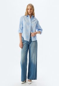 Camisa de botones azul claro con bordado floral, combinada con jeans de pierna ancha desgastados y zapatos blancos de punta. Tejido suave, ajuste casual.