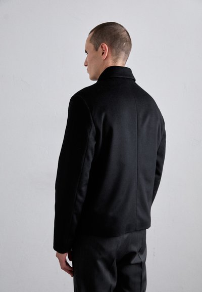 Black, fitted jacket with a collar, smooth texture, and minimal design. View from the back, paired with dark trousers.