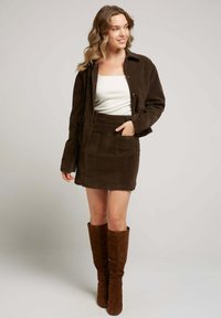 Femme avec des cheveux ondulés portant une veste en daim marron foncé, une jupe assortie avec des poches avant, un haut blanc et des bottes brunes jusqu'aux genoux, souriante.