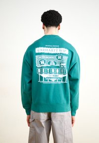 Turkusowy sweter z białym nadrukiem na plecach, z napisem "Detroit Super Market" i "Carhartt WIP" w grubych czcionkach. Klasyczny design z okrągłym dekoltem.