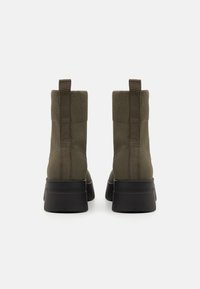 Botine verzi din material textil, cu glezna riblată, talpă neagră și tabă de prindere pe spate. Textură netedă și design structurat, potrivite pentru diverse ținute.