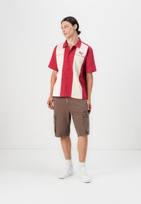 Rød og kremfarget kortermet skjorte med knappfront, med et brystlogo. Brune cargo-shorts med sidelommer, hvite sneakers.
