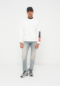 Uomo che indossa una felpa bianca Diesel con logo sulla manica, jeans chiari, cappellino nero e scarpe da corsa nero-bianco, in piedi su sfondo bianco.