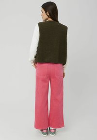 Gilet shaggy vert, chemise blanche à manches longues, pantalon en velours côtelé rose à jambes larges et baskets avec des accents gris et rose, vus de dos.