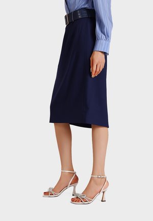 Donna che indossa una gonna blu navy al ginocchio, camicia a righe blu, cintura nera e sandali bianchi con tacco alto e cinturini con dettaglio a fiocco.