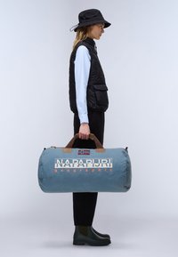 Borsa da viaggio blu con manici in pelle marrone, con logo e bandiera norvegese. Il modello indossa un gilet nero e stivali di gomma verdi.