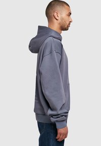 Grå hættetrøje med en frontlomme, oversized design og ribbede manchetter. Har snøre ved hætten og en blød tekstur, båret med blå jeans.