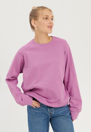 Νεαρή γυναίκα με ξανθά μαλλιά σε κότσο φοράει ένα φαρδύ λιλά φούτερ και μπλε τζιν, ποζάροντας με το ένα χέρι στη μέση.