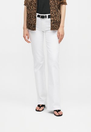 Žena oblečená v bielych rozšírených nohaviciach, čiernom páse, čiernom topu, košeli so vzorom leopardy a čiernych sandáloch s otvorenou špičkou, stojaca pred bielym pozadím.