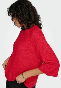 Rdeč pleteni pulover s tremi četrtinami rokavov, zaobljenim izrezom in mehko teksturo. Rob je rahlo ukrivljen, sproščen kroj.