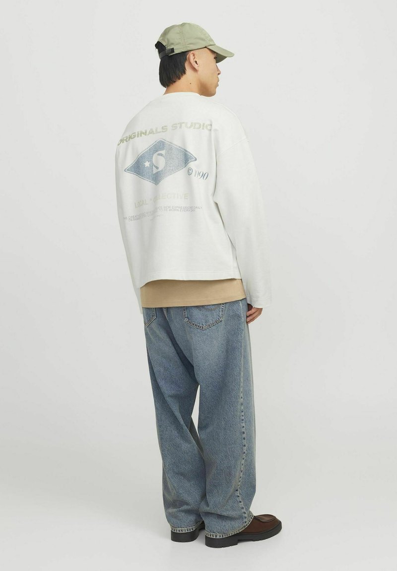 Sudadera blanca extragrande con print gráfico en la parte posterior, superpuesta a una camiseta beige. Llevada con unos jeans claros y holgados de color azul y botas marrones.