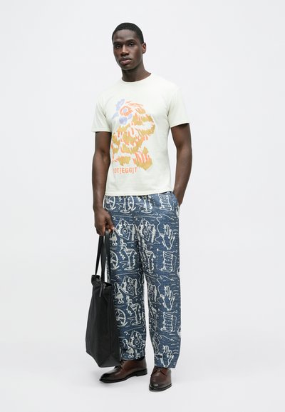 Ανοιχτό πράσινο βαμβακερό t-shirt με πολύχρωμο σχέδιο στο μπροστινό μέρος. Φαρδιά μπλε παντελόνια με σχέδια, μαύρη τσάντα tote και καφέ παπούτσια.