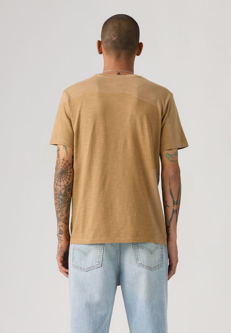 Man with short hair and arm tattoos wearing a plain brown t-shirt and light blue jeans, facing away against a plain background.