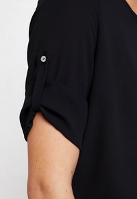 Musta paidan hiha, valmistettu sileästä kankaasta, jossa on napsautettavalla napilla kiinnitetty käännetty kalvosin. Malli on yksinkertainen ja istuva.