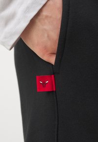 Sorte joggebukser med en glatt tekstur og et rødt merke med en okse-logo, plassert nær en lomme. Hvit skjorte mansjett synlig.