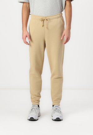 Uomo che indossa pantaloni da jogging beige con coulisse in vita e sneaker bianche ASICS, in piedi davanti a uno sfondo bianco semplice.