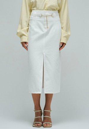 Mujer con falda lápiz midi blanca con abertura frontal y cinturón de cadena dorada, combinada con una blusa de manga larga color amarillo pálido y tacones beige con tiras.