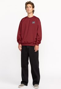 Sweatshirt bordeaux à col rond et poignets côtelés, associé à un pantalon large noir et des chaussures à enfiler à carreaux. Présente un petit logo brodé sur la poitrine.