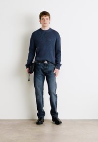 Maglione a coste blu navy, jeans blu scuro, scarpe nere e borsa nera piccola. Design semplice e dritto con una vestibilità rilassata su uno sfondo neutro.