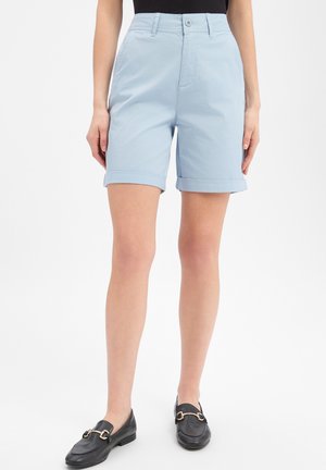 Vrouw draagt lichtblauwe omslagshort, zwart topje en zwarte loafers met gouden gespen, staand tegen een effen witte achtergrond.