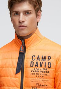 Orangefarbene gesteppte Jacke mit hohem Kragen, mit Reißverschluss. Enthält gedruckte Texte und Logodetails in Schwarz und Dunkelblau.