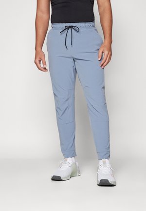 Homme portant un pantalon de jogging bleu clair avec un cordon noir et des chaussures de sport blanches, debout devant un fond uni.