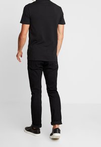 Hombre vestido con una camiseta polo negra, jeans negros arremangados y zapatillas negras con suelas blancas, de pie contra un fondo claro y sencillo.