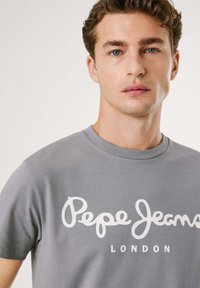 Šedé tričko s krátkým rukávem vyrobené z měkké bavlny, s výrazným bílým textem "Pepe Jeans LONDON" přes hruď v uvolněném, psacím písmu.