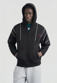 Νέος άνδρας που φοράει μαύρο φούτερ Adidas με φερμουάρ και λευκές ραφές, καθώς και ανοιχτό γκρι φόρμα Adidas, στέκεται με τα χέρια στις τσέπες.