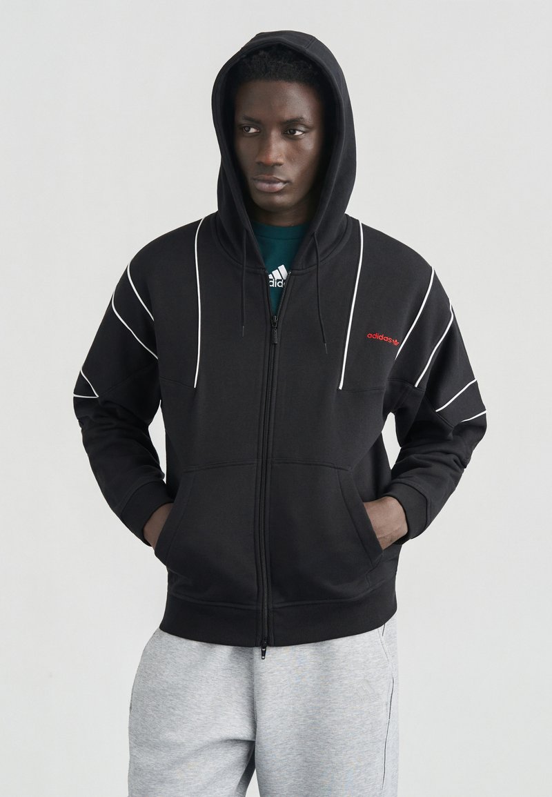 Νέος άνδρας που φοράει μαύρο φούτερ Adidas με φερμουάρ και λευκές ραφές, καθώς και ανοιχτό γκρι φόρμα Adidas, στέκεται με τα χέρια στις τσέπες.