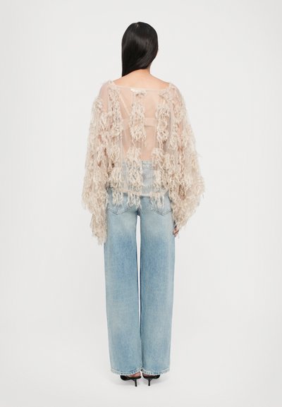 Durchsichtige, beige Bluse mit strukturierten Fransen, kombiniert mit hellblauen, weiten Jeans und schwarzen High Heels. Rückansicht des Outfits.