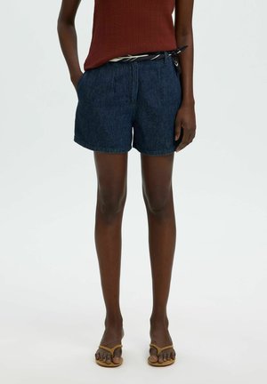 Personne portant un short en jean bleu foncé, un haut marron sans manches, une ceinture noire et blanche et des tongs beige, debout devant un fond uni.
