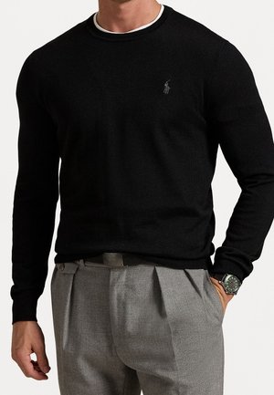 Mand iført sort langærmet sweater med logo, grå plisserede bukser, sort bælte og armbåndsur, stående med den ene hånd i lommen.