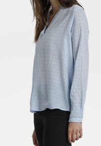 Eine Frau trägt eine hellblaue Bluse mit langen Ärmeln und weißen Blattmustern sowie schwarze Hose und steht vor einem schlichten weißen Hintergrund.