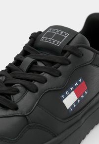 Crne kožne tenisice s zaobljenim vrhom, debelim potplatom, crnim vezicama i istaknutim Tommy Jeans logotipom u crvenoj, bijeloj i plavoj boji.