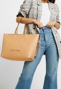 Kožená taška v opálené barvě s embosovaným nápisem "NY" a "Calvin Klein". Dvoje ramenní popruhy a strukturovaný design doplňují ležérní outfit.