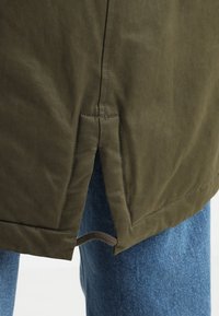 Veste vert olive à finition texturée, avec une fente latérale à l'ourlet et un détail à cordon. Portée avec un jean en denim bleu clair.