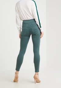 Jeansy w kolorze niebiesko-zielonym o wysokim stanie, dopasowanym kroju, elastycznym materiale i wyszywanymi tylnymi kieszeniami. Połączone z białą bluzką z długim rękawem.